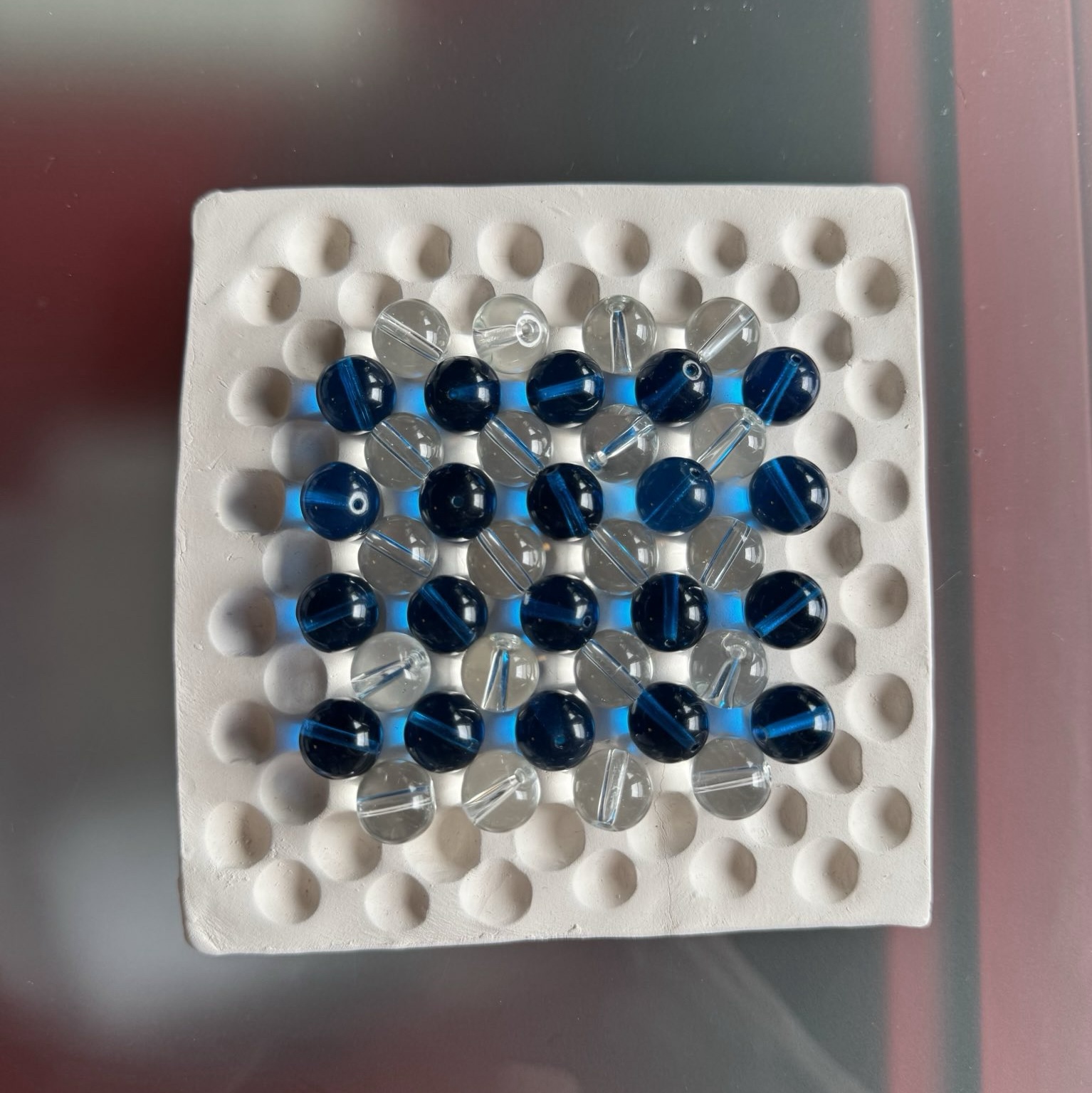 flat ceramic square with grid of divets and blue and transparent beads resting on top